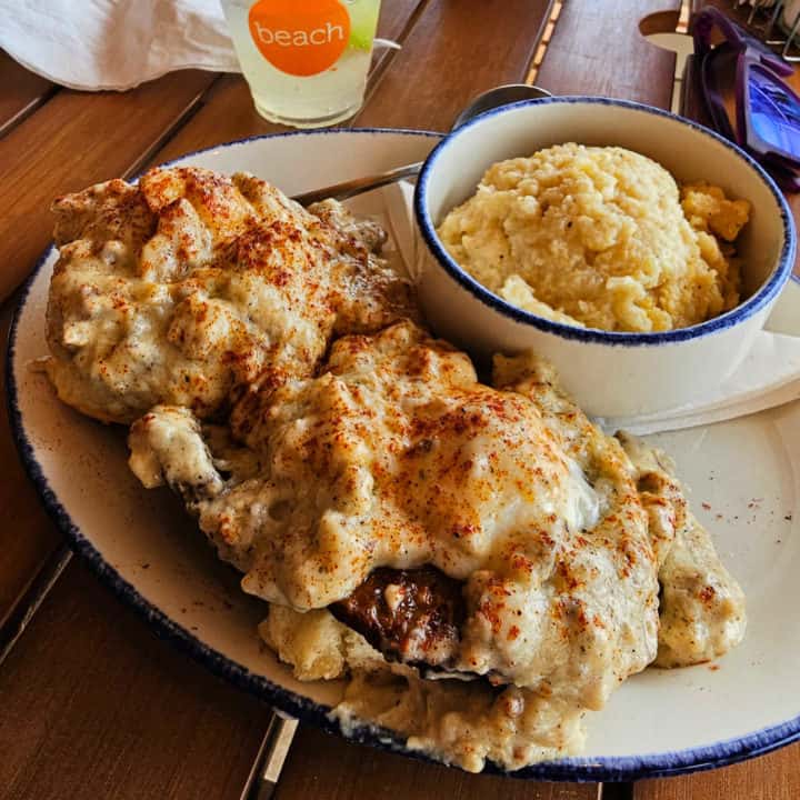 Sausage gravy covered biscuits next to a bowl of grits and a Orange Beach glass.