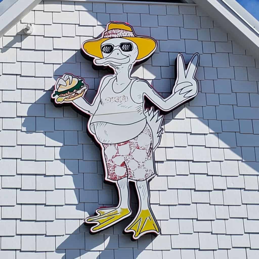 Cartoon duck with a yellow hat, holding a burger, and giving the piece sign on the side of a building.