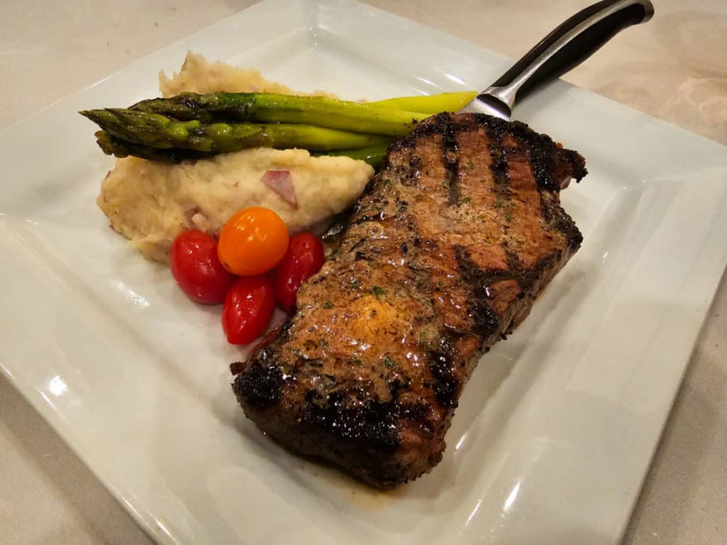 Angus steak on a white plate next to potatoes, tomatoes, asparagus and a knife