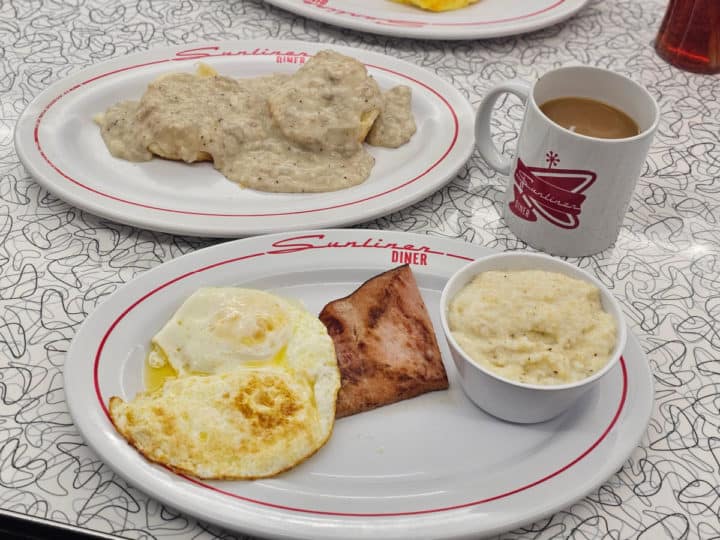 biscuits and gravy next to a plate with ham, eggs, and grits and a Sunliner Diner Mug