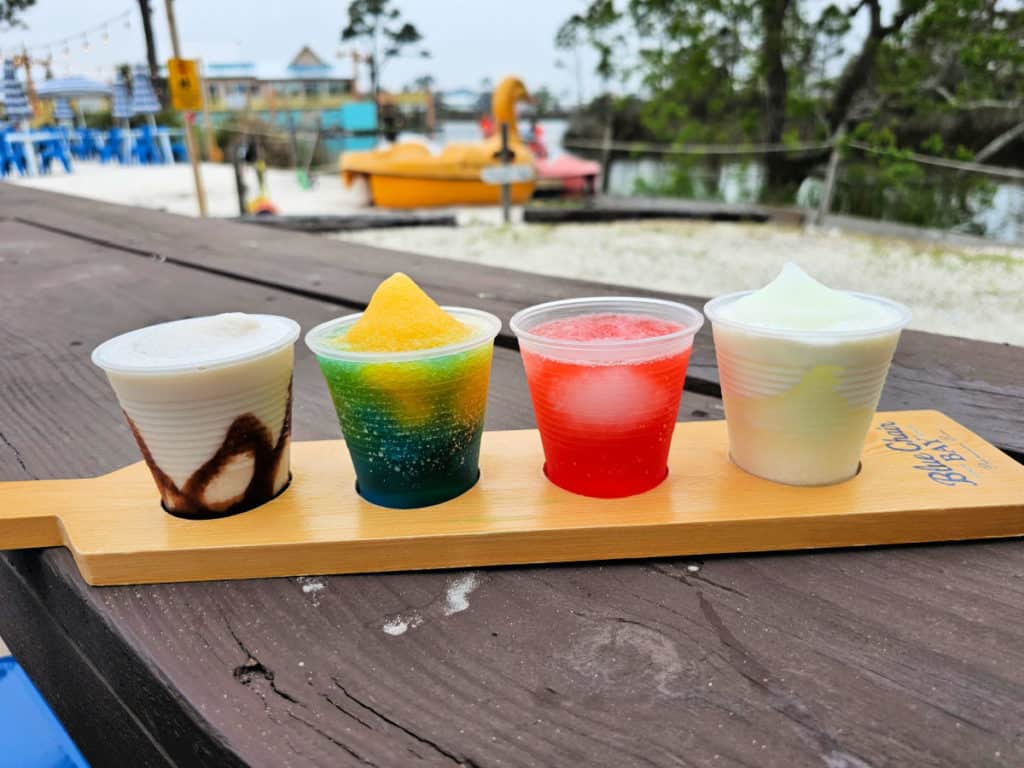 Four frozen multi colored daiquiris on a wooden board with a yellow duck pedal boat in the background