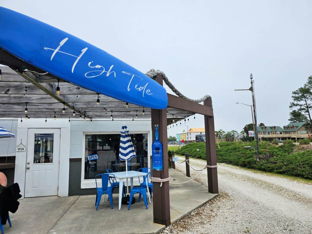 High Tide printed on a blue surfboard over a walkway leading to a white door, patio with blue chairs, white tables and umbrellas. 