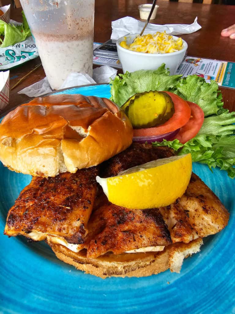 fish sandwich with lemon, pickles, tomato, and lettuce on a blue plate next to a bushwacker and side dish