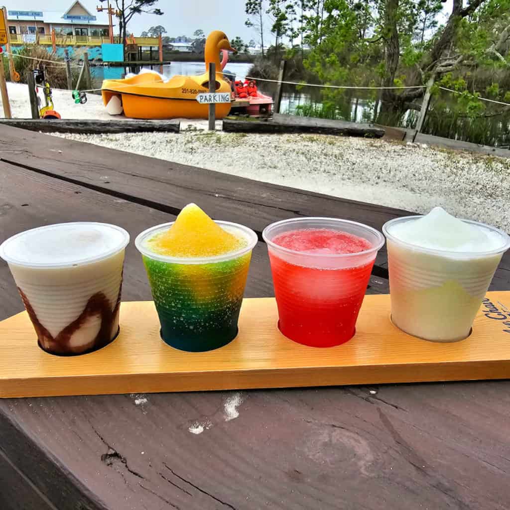 High Tide Daiquiri sampler on a wooden board, sitting on a outdoor table with views of the water and a yellow duck paddle boat