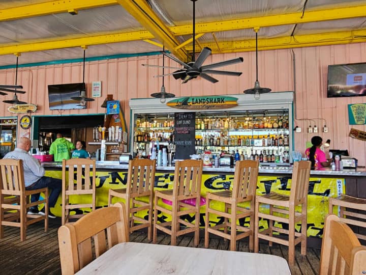 indoor bar with tall chairs next to the bar, Landshark surfboard, fans over head with a person sitting at the bar. 