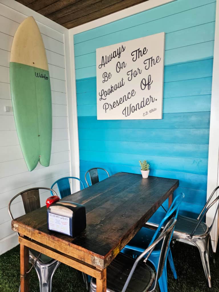 Table and chairs next to a turquoise wall with a quote and a surfboard