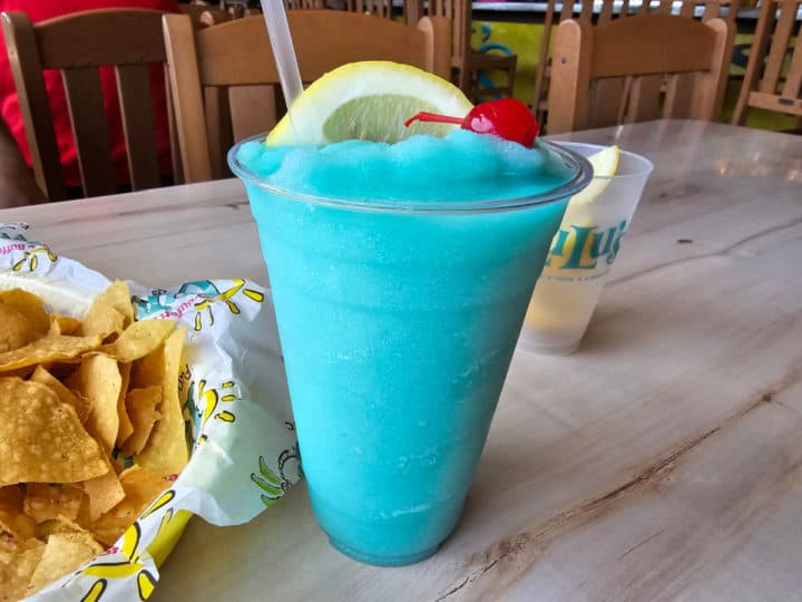 Blue frozen cocktail in a plastic cup with a lemon wedge and cherry next to a LuLu's cup and basket of chips