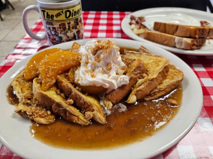 French Toast with whipped cream and peach slices on a white plate next to a Ugly Diner mug