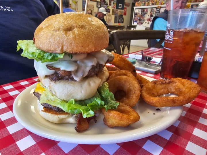 Large multi layer burger with cheese, bacon, and lettuce next to onion rings and an Iced Tea on a checkered tablecloth