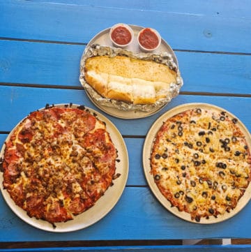 Surfside Pizza garlic bread with marinara sauce, meat pizza, and pineapple black olive pizza on a blue picnic table