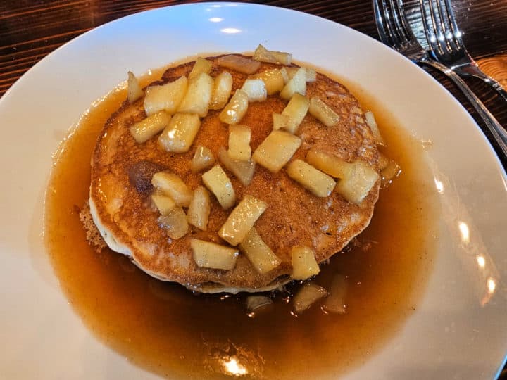 Stack of pancakes covered in apple pieces and syrup on a white plate
