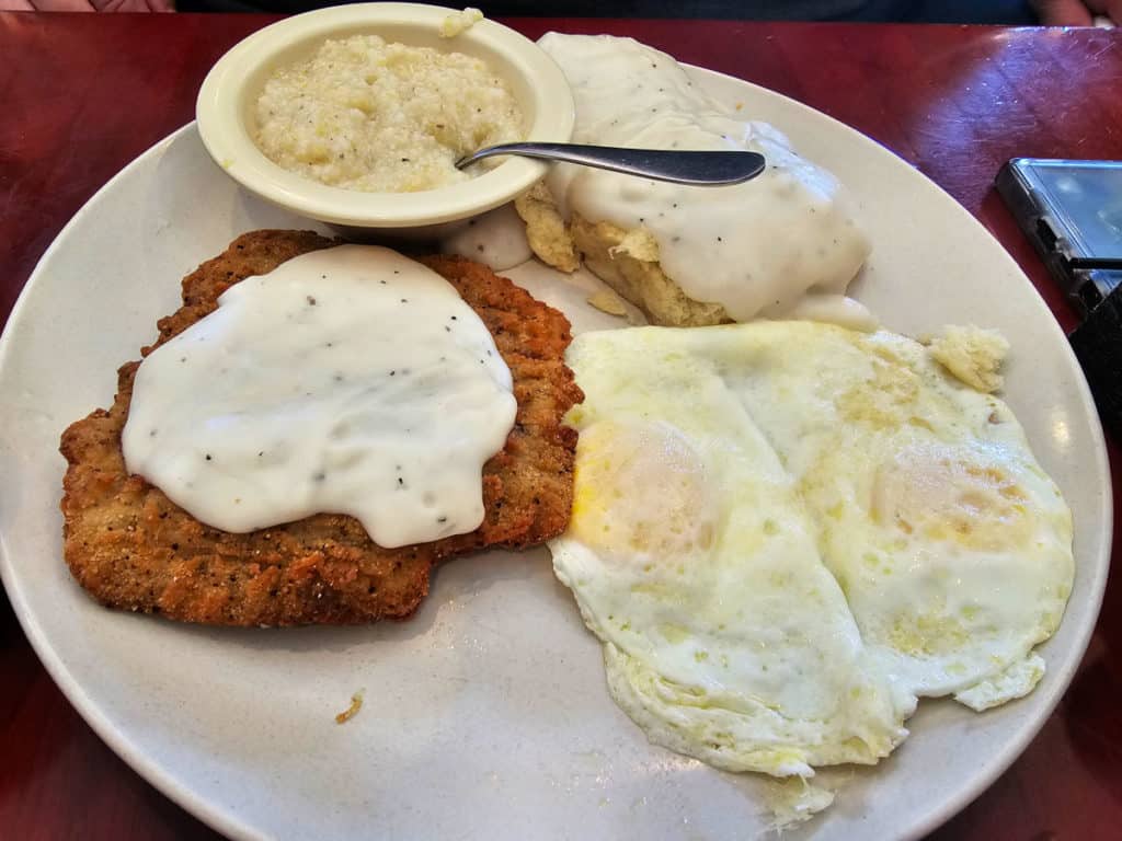 Country fried steak with gravy, grits, biscuits and gravy, and two eggs on a white plate