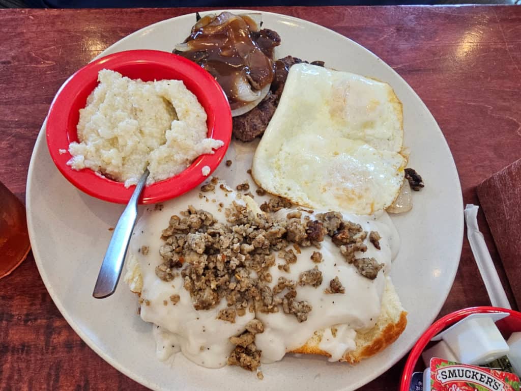 smothered hamburger steak, grits, over medium eggs, and sausage gravy and biscuits on a white plate
