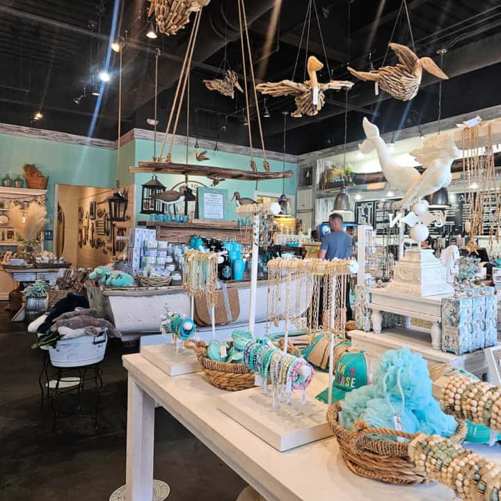 Coastal gift shop with pelicans made with driftwood hanging from the ceiling, tables with jewelry, hats, and gifts, bird statues, and a coffee bar