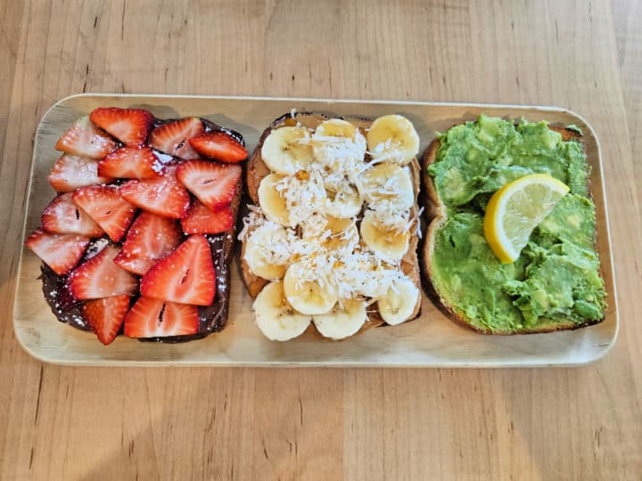 Toast flight with three slices of toast, strawberry and nutella, banana coconut peanut butter, and avocado toast on a platter