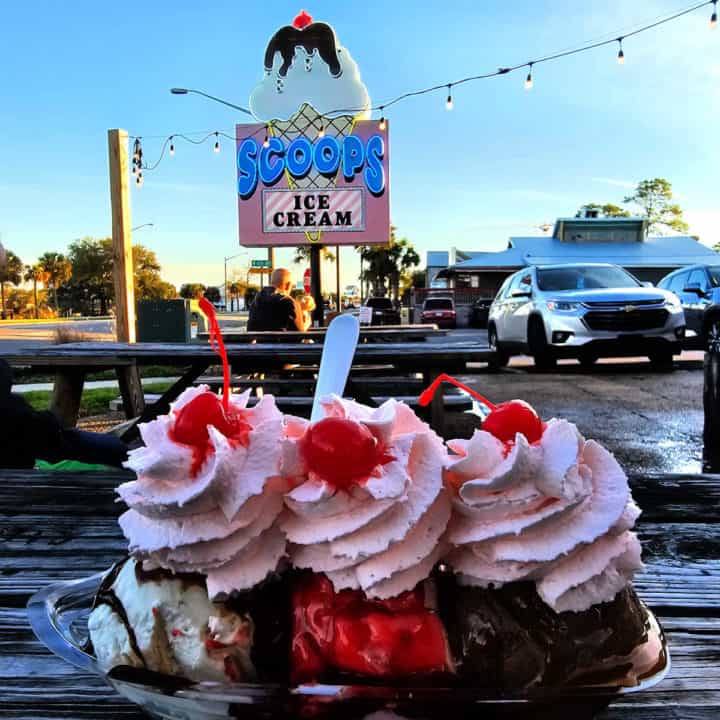 banana split with pink whipped cream and cherries on a picnic table with Scoops Ice cream sign in the background 