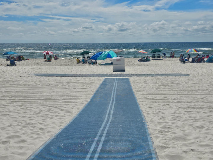Blue wheelchair mat leading onto the sand with umbrellas and people near the water. 