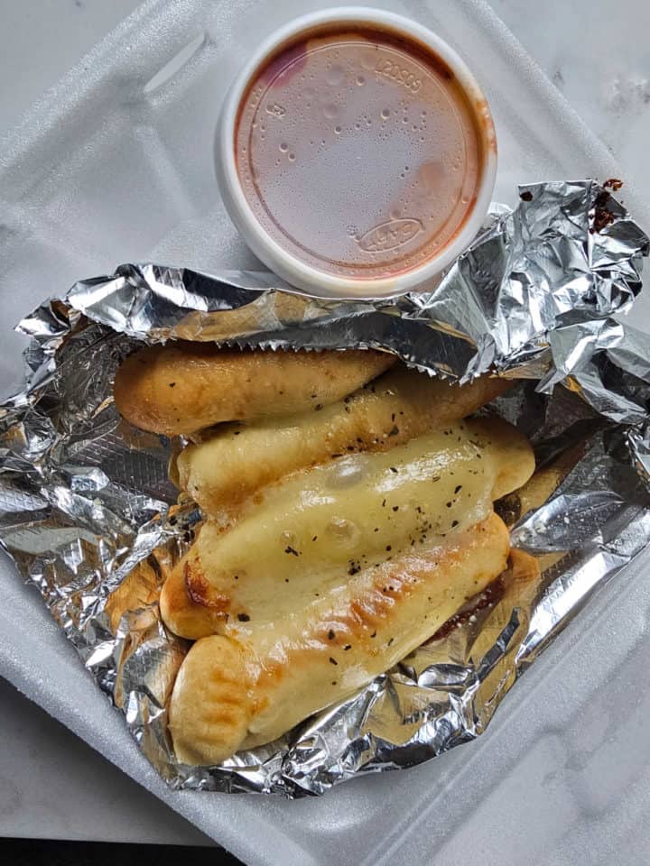 Cheesy breadsticks wrapped in foil in a to go container with marinara sauce 