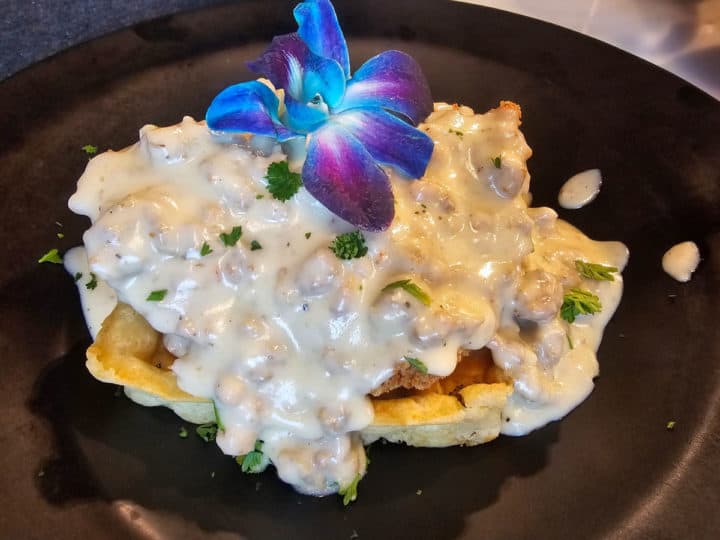 gravy covering a waffle and chicken on a dark plate with an orchid on top