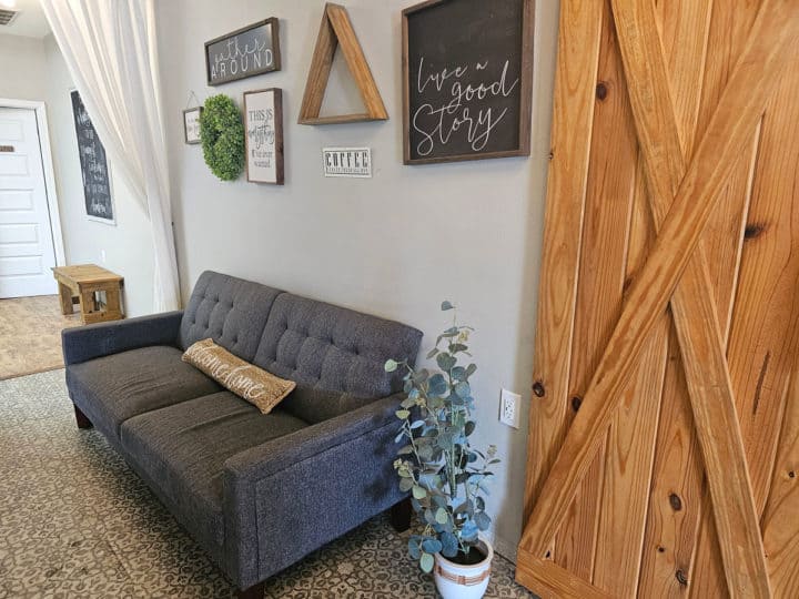 Grey couch with a welcome home pillow below signs on the wall next to a wooden door