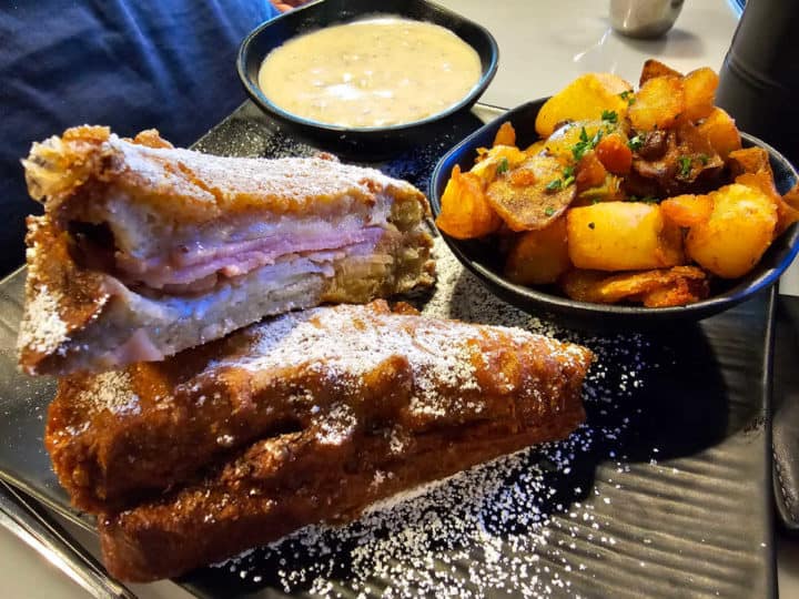 Monte Cristo sandwich on a dark plate with powdered sugar next to a bowl of gravy and a bowl of breakfast potatoes