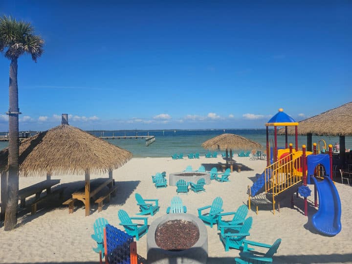 Adirondack chairs around a fire pit, kids play area, and thatched huts on the beach