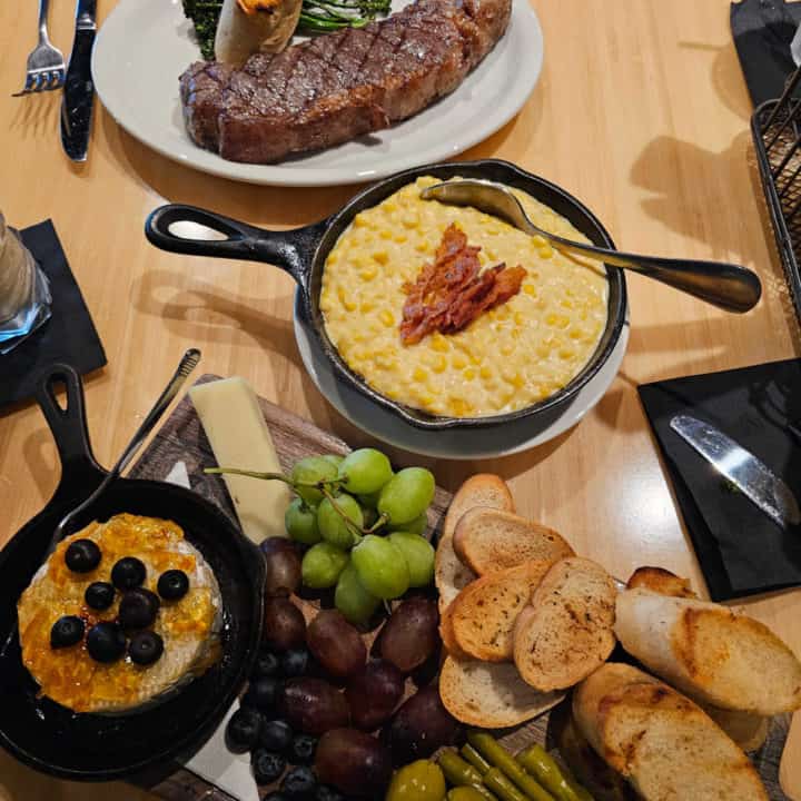 wood table with a steak dinner, corn in a cast iron skillet, baked brie on a cutting board with bread and grapes