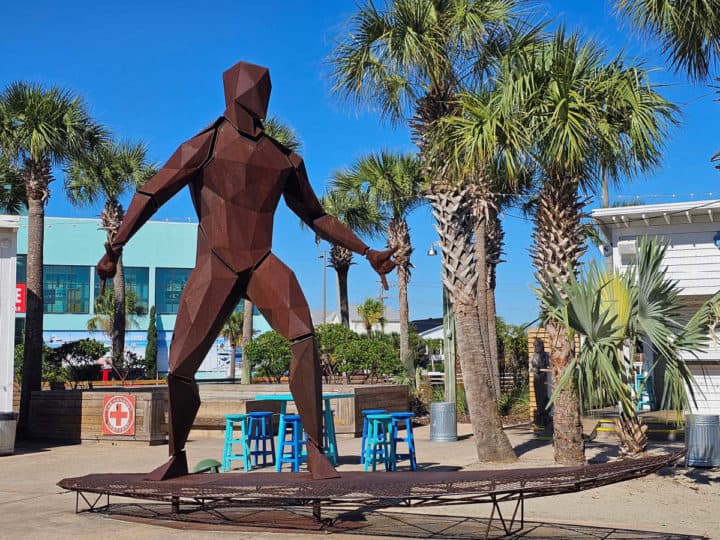 Larger metal surfer statue on a surfboard near palm trees