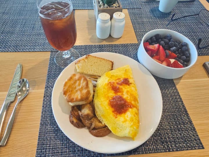 omelet, potatoes, biscuit, and lemon loaf on a white plate next to a bowl of fruit and glass of iced tea