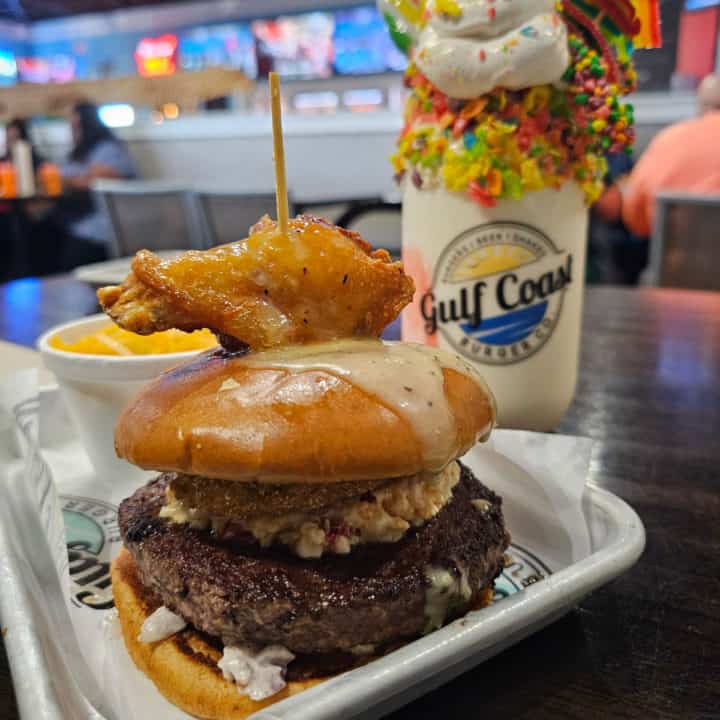 Burger with a chicken wing on the top in front of a large shake in a Gulf Coast Burger Co glass topped with whipped cream and fruity pebbles. 