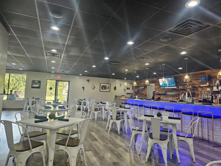 tables and chairs inside the restaurant with a bar with stools and tvs in the background. 