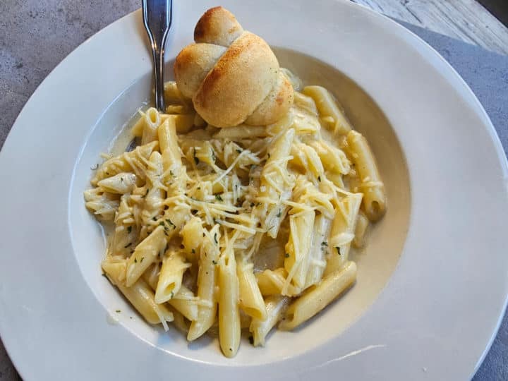 Penne noodles with alfredo sauce in a white bowl with a knot roll