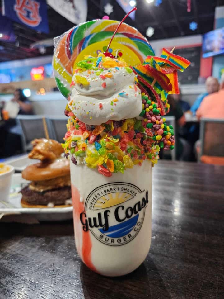 Gulf Coast Burger Co glass with a giant milkshake covered in whipped cream, fruity pebbles, a sucker, and candy