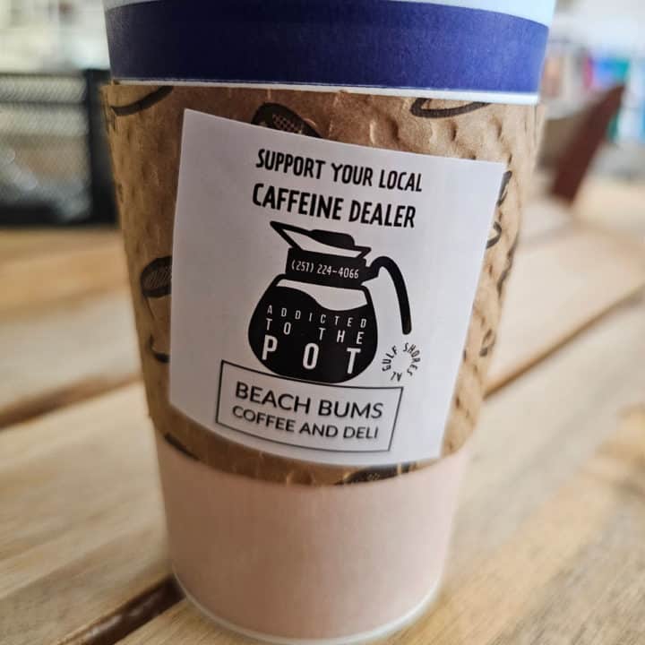 Paper coffee cup with a sticker that says Support your local caffeine dealer, Beach Bums Coffee and Deli