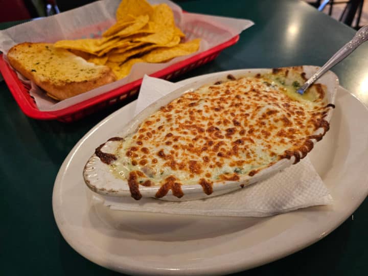 spinach artichoke dip in a white bowl next to a basket of chips and garlic bread