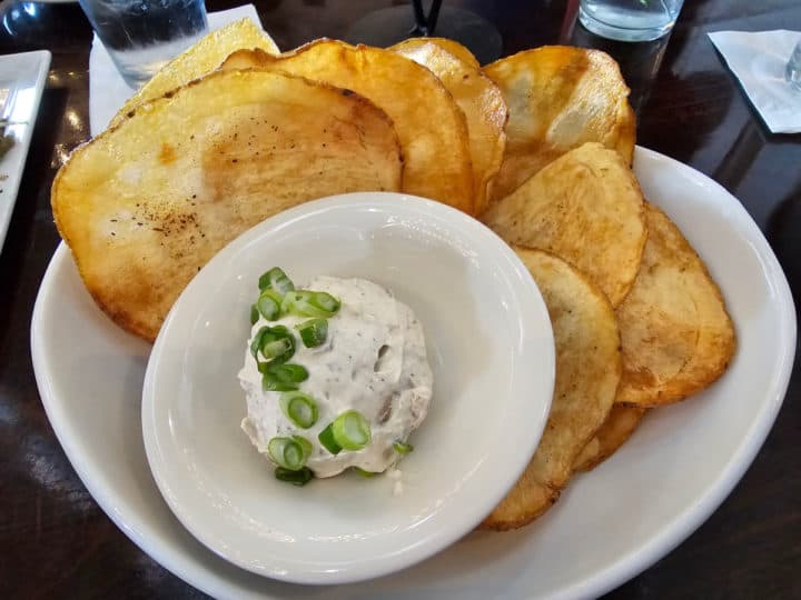 Large potato chips on a white plate next to a bowl with dip covered in green onions 