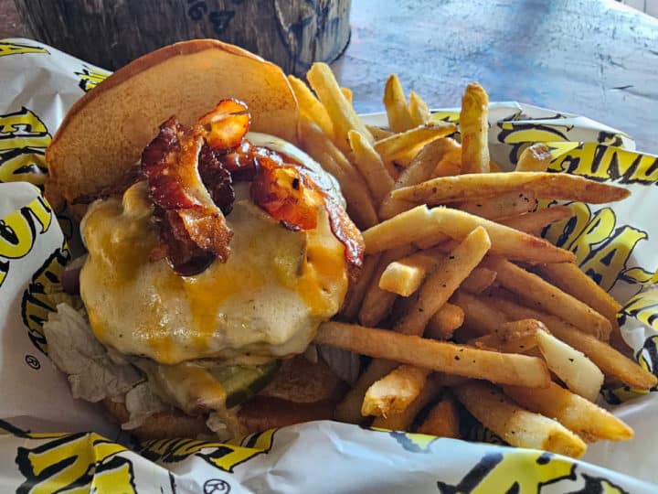 Burger with cheese and bacon next to fries in a Flora Bama paper wrapped basket