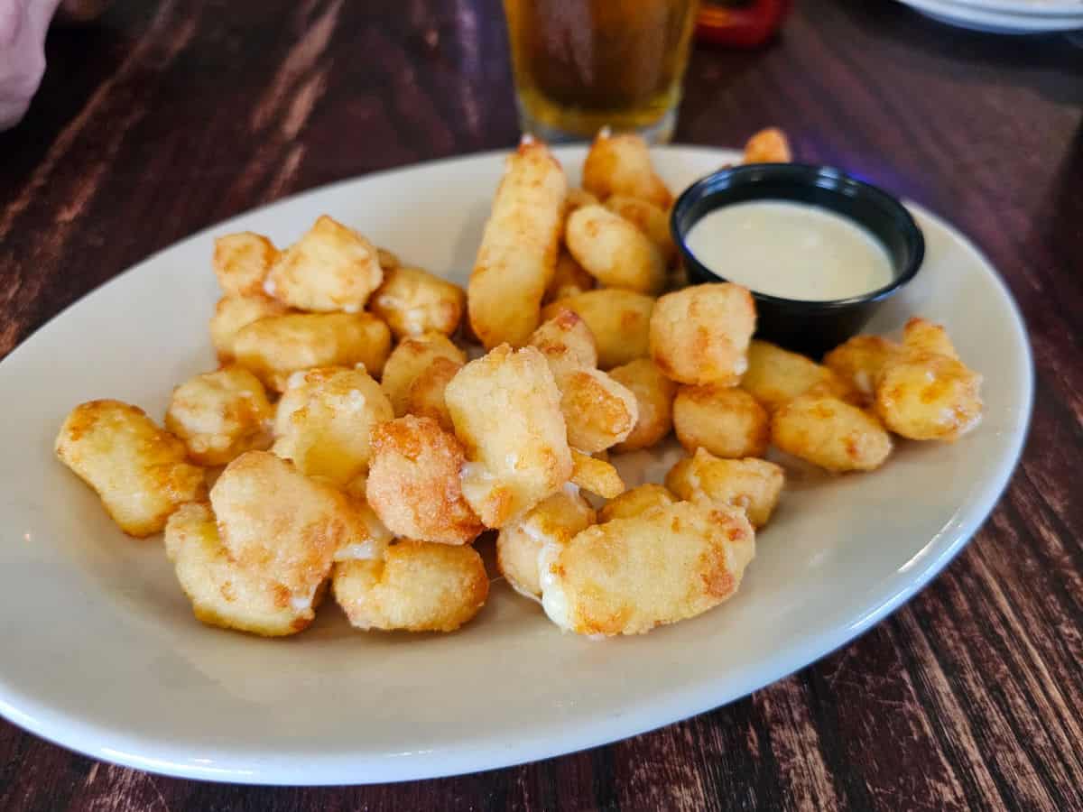 Cheese curds on a white plate wtih a small container of ranch next to a glass of beer. 