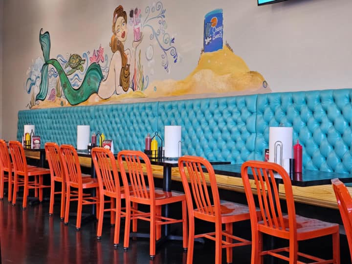 Mermaid painting on the wall above a long bench with a long table and orange chairs 