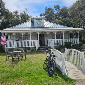 Exterior of Lighthouse Bakery with a white wooden bridge leading over the grass, a bike against the bridge, and tables in the lawn.
