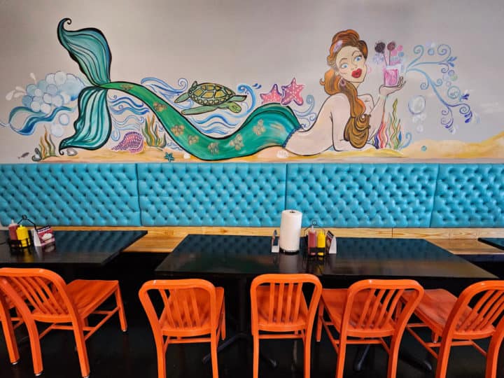 Mermaid painting with turquoise tail holding a milkshake over a long table with orange chairs. 