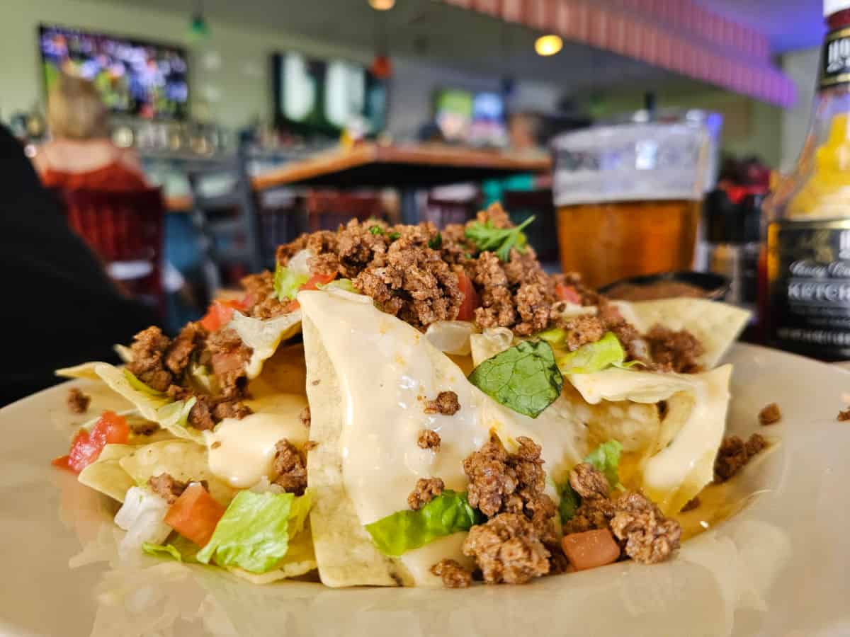 Nachos with ground beef next to a beer on a table with TVs and people in the background