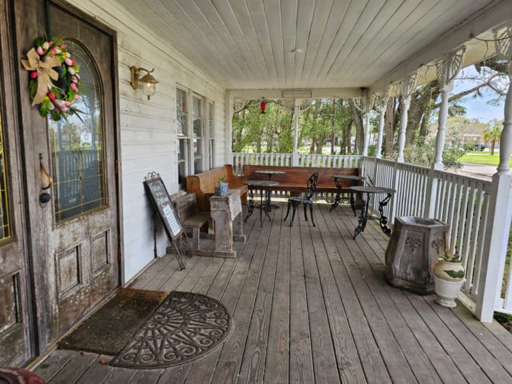 Outdoor seating on a large front porch with benches, and small tables next to a wooden door. 