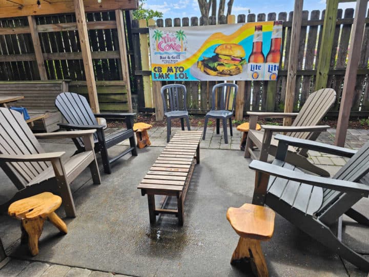 Outdoor seating with a wooden thin table next to Adirondack chairs, regular chairs, and stools with a Cove poster in the background