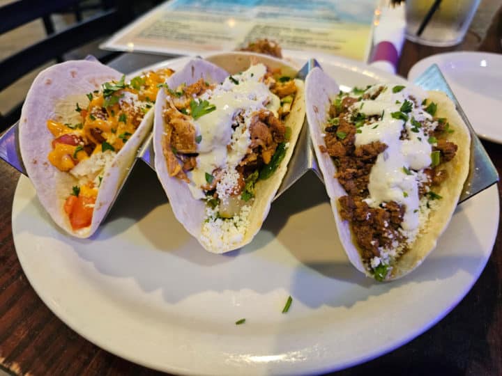 three tacos on a white plate with a menu in the background