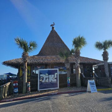 The Tiki at Sportsman Harbor sign in front of a thatched covered bar with a pelican on top of it and palm trees
