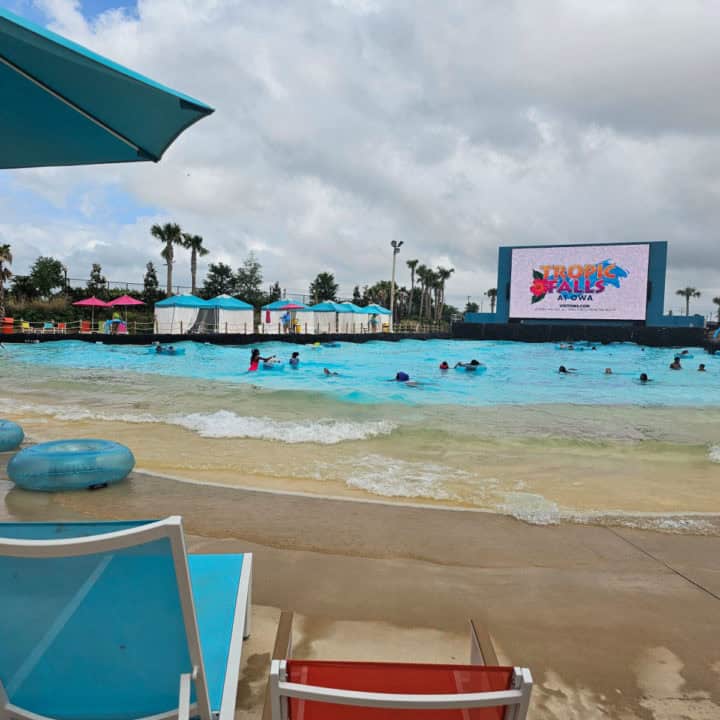 Large wave pool with a Tropic Falls logo on a large screen, turquoise lounge chair, and innertube near the shore