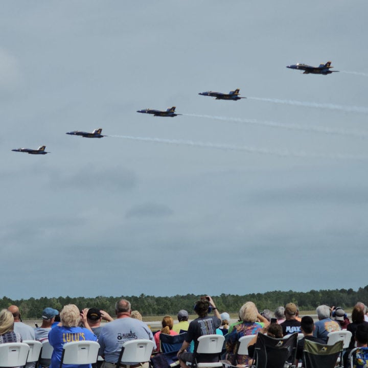 Five Blue Angels jets fly over a crowd sitting in chairs along the flight line