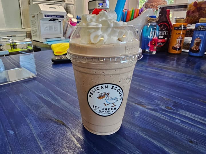 Plastic cup with Pelican Scoops Ice Cream sticker on the front sitting on a counter with ice cream supplies behind it. 