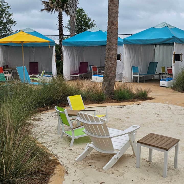 Three cabanas with blue roofs seen over beach chairs in a sandy area. 